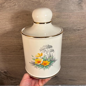 Può includere: Un barattolo di ceramica bianca con un bordo dorato e un motivo floreale con fiori gialli e verdi. Il barattolo ha un coperchio con la parte superiore arrotondata.