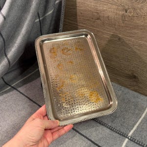 Könnte beinhalten: Ein rechteckiges silbernes Tablett mit einem Perlenrand und einer strukturierten Oberfläche. Das Tablett hat einige Flecken auf der Innenseite. Das Tablett wird von einer Person gehalten.