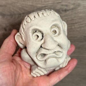 May include: A small, off-white, sculpted figurine of a grumpy-looking face with large eyes, a prominent nose, and a furrowed brow. The figure appears to be resting on its hands. The sculpture is held in a person's hand.