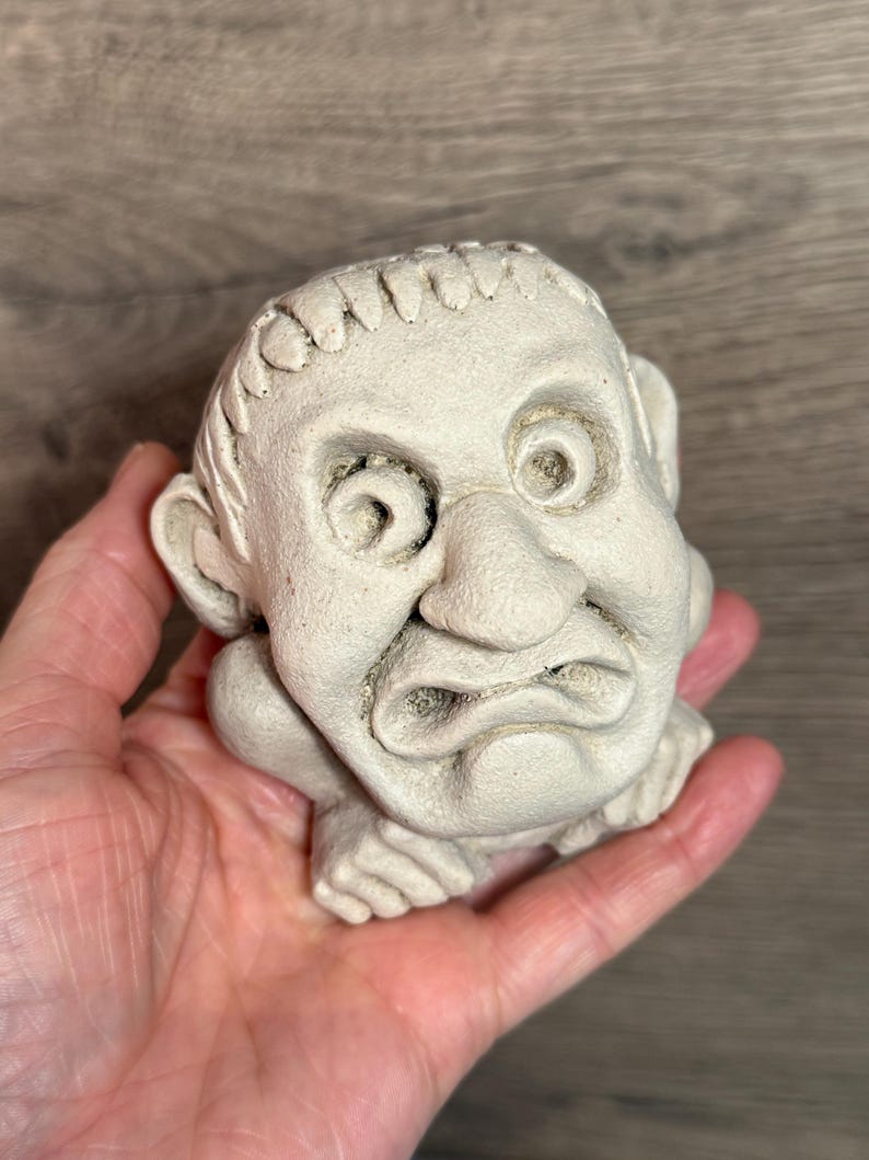May include: A small, off-white, sculpted figurine of a grumpy-looking face with large eyes, a prominent nose, and a furrowed brow. The figure appears to be resting on its hands. The sculpture is held in a person's hand.