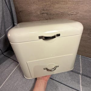 Puede incluir: Panera metálica de color crema con tapa curvada y cajón desplegable. La caja tiene un asa plateada en la parte superior y un asa a juego en el cajón. Un artículo de almacenamiento de cocina.