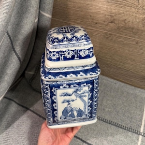 May include: Blue and white ceramic jar with a square shape and a lid. The jar features detailed illustrations of figures and landscapes, with floral and geometric patterns. The jar is held in a hand.