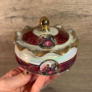 Puede incluir: Una caja de porcelana para alhajas con una tapa dorada y un borde festoneado. La caja está decorada con un patrón floral rojo y dorado y una escena de dos personas bailando. La caja tiene tres pequeños pies.