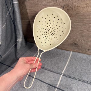 Op de afbeelding: Crèmekleurige keukenzeef met een lange handgreep. De zeef heeft een afgeronde vorm met kleine, gelijkmatig verdeelde gaatjes. De handgreep is ook crèmekleurig en lijkt van metaal te zijn. Ontworpen voor het zeven van voedsel, zoals pasta of groenten.