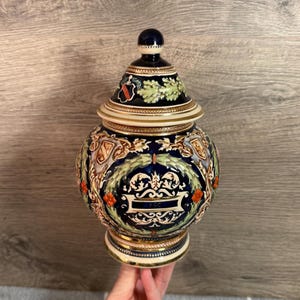 Op de afbeelding: Een decoratieve keramische pot met een deksel. De pot is voornamelijk marineblauw met gouden randen en heeft een gedetailleerd ontwerp van wapens, bloemmotieven en een centraal cartouche. Het deksel is bekroond met een donkerblauwe knop.