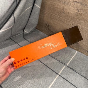 May include: An orange knitting needle gauge with the words "Knitting Needles" in cursive script. The gauge has multiple holes of varying sizes and a dark brown cap. The gauge is on a gray and white plaid blanket.