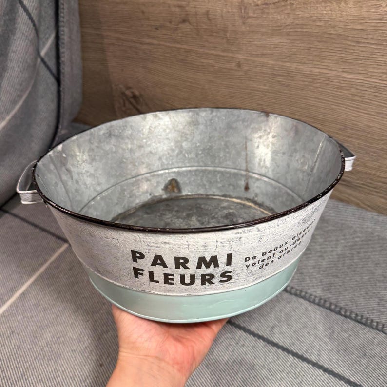 Op de afbeelding: Een ronde kom van gegalvaniseerd metaal met twee handvatten aan de zijkanten. De buitenkant heeft een witte band met de zwarte tekst PARMI FLEURS en kleinere tekst, terwijl het onderste gedeelte is geverfd in lichtblauw met een verweerde look.
