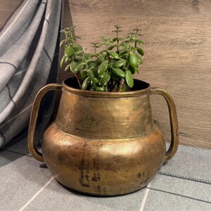 May include: A weathered brass planter with two side handles containing a green succulent plant. The metal vessel features a rounded base and a slightly tapered neck, showing a tarnished, aged patina across its entire exterior surface.