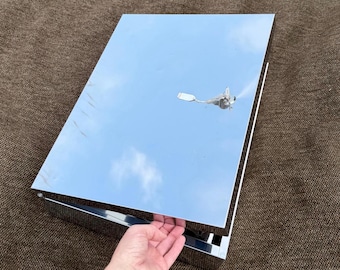 Op de afbeelding: Een rechthoekige doos van gepolijst chroom met een spiegelend oppervlak. Het deksel staat gedeeltelijk open en onthult een donkere binnenkant. De buitenkant is sterk reflecterend en toont een reflectie van de lucht en een kleine vogel.