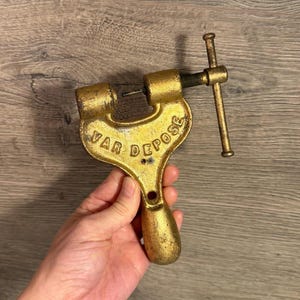 May include: A vintage gold-toned metal hand tool with a C-shaped frame and a threaded screw mechanism. The tool features the embossed text VAR DEPOSE on the front surface. It has a rounded handle at the bottom and a T-shaped handle at the top.