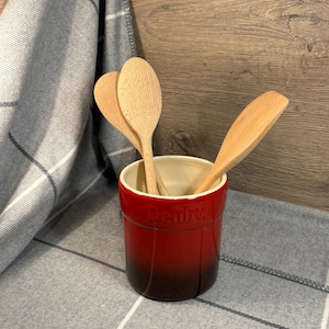 May include: Red ceramic utensil holder with the word "Denby" printed on the side. The holder is filled with wooden spoons.