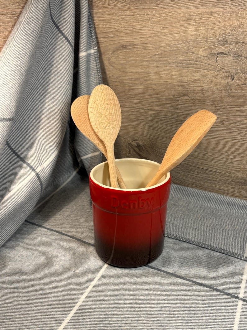 May include: Red ceramic utensil holder with the word "Denby" printed on the side. The holder is filled with wooden spoons.