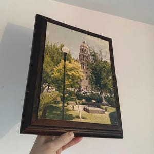 Puede incluir: Fotografía enmarcada de una iglesia con una alta torre campanario, rodeada de árboles y un parque. El marco es de color marrón oscuro y la imagen tiene una estética vintage. Una farola es visible en primer plano.