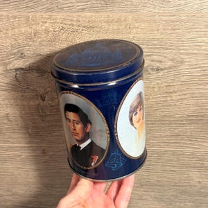 May include: A vintage blue tin featuring portraits of Charles and Diana. The tin has a domed lid with an architectural design. The portraits are in oval frames. The tin is held by a hand.