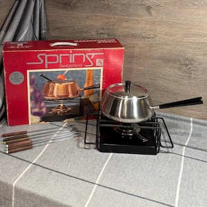 Può includere: Un set per fondue con una pentola in acciaio inossidabile, un supporto in metallo nero con un bruciatore e sei forchette per fondue. Il set è confezionato in una scatola rossa con la scritta "Springs" e "Svizzera" stampata su di essa.