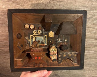 May include: A wooden shadow box diorama depicting a rustic kitchen scene. The interior features a carved figure seated near a stove, a table, and a window with a painted landscape. The frame is dark with a black border.