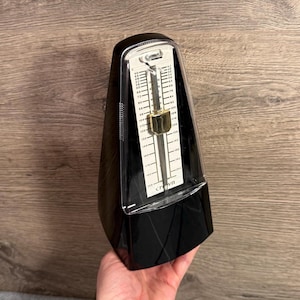 May include: A black Crown metronome with a clear plastic cover. The metronome has a pendulum with a gold-coloured weight and a white scale with tempo markings. The base is black and angled.