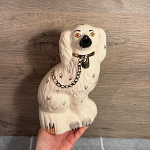 Puede incluir: Figura de perro de cerámica blanca con detalles dorados y negros. El perro está representado en posición sentada, con un collar y una cadena dorados. Los ojos son naranjas y la nariz negra. La figura está sujeta por una mano.