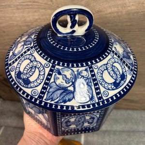 Op de afbeelding: Blauwe en witte keramische pot met deksel en een knopje in de vorm van een pretzel. De pot heeft een zeshoekige vorm en is versierd met een herhalend patroon van pretzels, bladeren en cirkels.