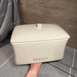 May include: A rectangular, off-white ceramic bread box with a matching lid featuring a small knob handle. The front of the container displays the word BREAD in a simple, dark, sans-serif font. The surface has a smooth, glossy finish.