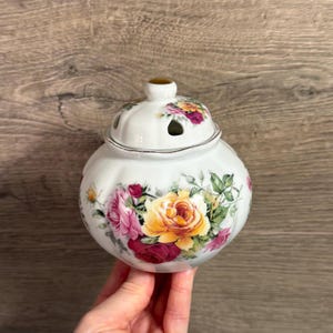 May include: A white ceramic jar with a lid, decorated with a floral pattern of yellow, pink, and red roses. The jar has a rounded shape and a small, round knob on top. The lid has several small holes.