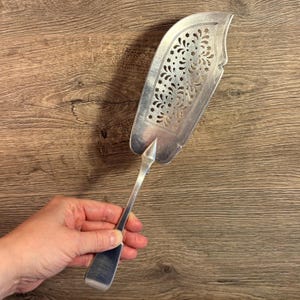 May include: A silver fish server with a decorative, pierced design. The server has a long handle and a wide, slotted blade with a floral and star pattern. The server is held in a hand against a wooden background.