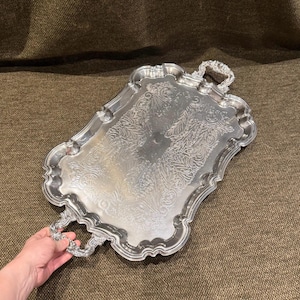 May include: A rectangular silver-toned serving tray featuring an ornate, embossed floral pattern across its surface. The tray has a scalloped edge and two decorative handles on opposite ends, with a polished finish reflecting light across the metal.