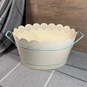 May include: An oval metal basin in cream with a scalloped, perforated rim featuring a floral pattern. The basin includes two light blue side handles and a thin light blue horizontal band encircling the exterior body of the container.