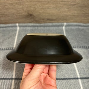 May include: A black ceramic bowl with a wide, flat rim. The bowl is held in a hand, showcasing its glossy finish and contrasting white interior. The bowl's design is simple and modern, suitable for serving various dishes.