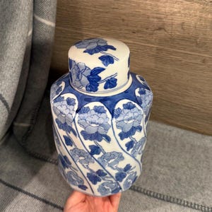 Puede incluir: Un jarrón de cerámica azul y blanco con tapa. El jarrón está decorado con un estampado floral en tonos azules y blancos. El jarrón está sostenido por una persona.