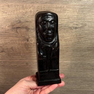 May include: A black wooden sculpture of a person wearing a hooded robe. The sculpture is carved in a simple style and has a minimalist design.