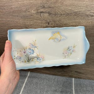 May include: A white rectangular serving tray with a blue border and a scalloped edge. The tray features a hand-painted design of blue and pink flowers and a yellow bird.