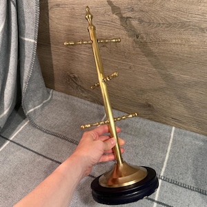 May include: A gold-colored metal mug tree with a black base. The tree has multiple arms to hold mugs and a decorative finial at the top. The mug tree is displayed against a wood-grain background.