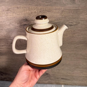 May include: A ceramic teapot with a speckled cream-coloured body, featuring a dark brown band around the base and a matching dark brown ring on the lid. The teapot has a curved handle on the left side and a short, angled spout on the right side.