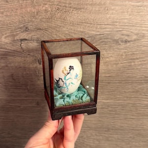 May include: A decorative egg with a painted Asian-inspired design, displayed inside a small, wooden-framed glass box. The egg rests on a bed of teal fabric. The box has a dark brown finish.