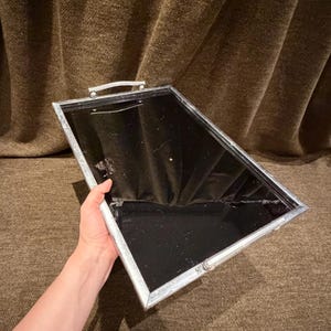 May include: A rectangular serving tray featuring a reflective black mirrored base and a metallic silver frame. The tray includes two silver handles attached to the shorter sides, providing a sleek and functional design for carrying items.