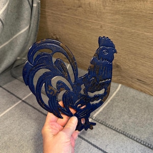 Pode incluir: Um descanso de panela em metal azul em forma de galo com acabamento envelhecido. O descanso de panela tem um design detalhado, incluindo crista, barbela e penas da cauda. Mede aproximadamente 20 cm de altura e 18 cm de largura.