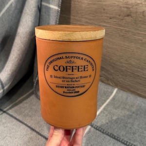 May include: A cylindrical, orange-brown ceramic coffee canister with a wooden lid. The canister features the text "COFFEE" in a decorative font, along with the words "The Original Suffolk Canister" and "Henry Watson Pottery, England, Founded 1800."