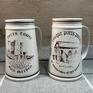 Op de afbeelding: Twee witte keramische bierpullen met zwarte en bruine accenten. Eén pul heeft een tekening van een kasteel en de tekst "Ypres Tower Gun Garden". De andere heeft een tekening van een kerk en de tekst "Cinque Ports Pottery the Monastery".