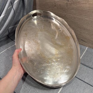 May include: Oval silver serving tray with an ornate floral and scrollwork design. The tray has a decorative edge and a handle on one side. The tray is held by a person's hand.