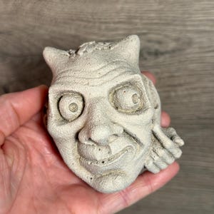 May include: A small, off-white, sculpted face with horns and a mischievous expression. The detailed carving features large eyes, a prominent nose, and a slight grin. The sculpture has a textured surface and is held in a hand.