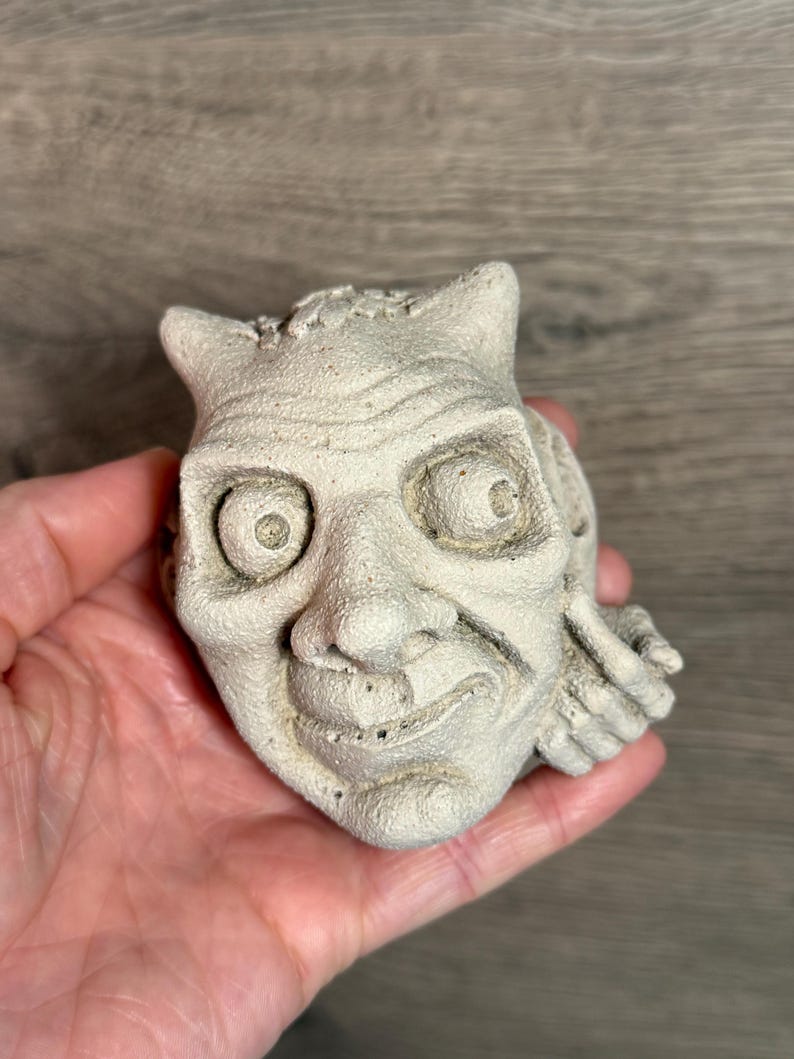 May include: A small, off-white, sculpted face with horns and a mischievous expression. The detailed carving features large eyes, a prominent nose, and a slight grin. The sculpture has a textured surface and is held in a hand.
