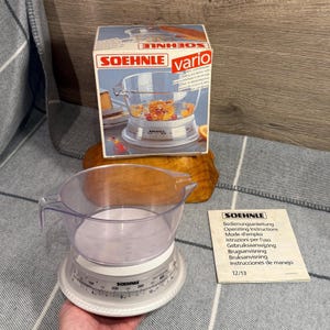 May include: A clear plastic SOEHNLE vario kitchen scale with a measuring cup, and a box with the same design. The scale has a white base with measurements in grams. An instruction manual is also present.