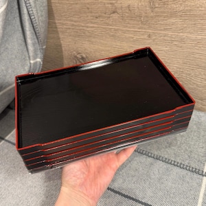 Puede incluir: Una pila de bandejas lacadas en negro con ribete rojo. Las bandejas rectangulares parecen ser de madera. Las bandejas están apiladas una encima de otra.