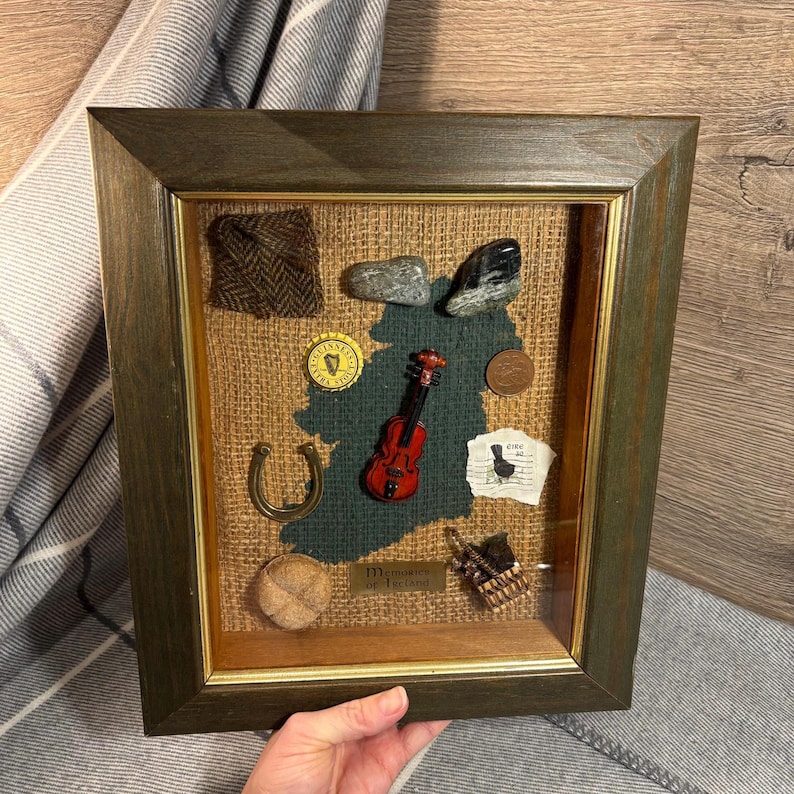 Puede incluir: Una caja de sombra enmarcada con un contorno verde de Irlanda, que presenta varios art&iacute;culos de tem&aacute;tica irlandesa, como un viol&iacute;n, una herradura, una muestra de tela de tweed, una piedra y una moneda. El texto "Memories of Ireland" es visible en una peque&ntilde;a placa.