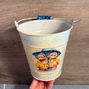 Pode incluir: Um pequeno balde de metal branco com uma alça e um detalhe azul. O balde apresenta uma ilustração de dois patinhos usando capas de chuva amarelas sob um guarda-chuva. O balde é segurado por uma pessoa.