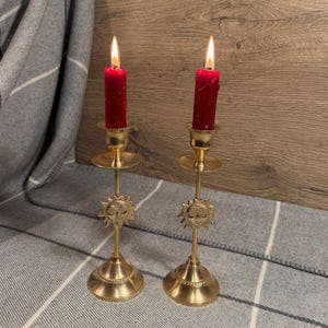 Puede incluir: Dos velas rojas encendidas en candelabros dorados ornamentados. Los candelabros tienen un diseño de cara de sol y están colocados sobre una superficie estampada gris y blanca. El fondo es una superficie de madera.