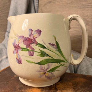 Puede incluir: Una jarra vintage de color crema con un diseño floral con iris púrpuras y verdes. La jarra tiene un asa dorada.