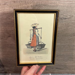 May include: Framed vintage print depicting a woman carrying baskets of strawberries. The woman wears a hat and apron, with the text "Greet a little Cabaren. Strawberries large and small" at the bottom. The frame is black with gold trim.