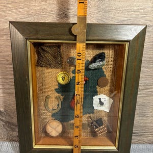 Puede incluir: Una caja de sombra enmarcada con un fondo de arpillera y varios art&iacute;culos de tem&aacute;tica irlandesa, incluyendo una herradura, un tr&eacute;bol, un peque&ntilde;o viol&iacute;n, una pelota de f&uacute;tbol, un trozo de tela de tweed, una peque&ntilde;a cesta, una moneda y un trozo de papel con el texto "C&iacute;rc", que en irland&eacute;s significa "c&iacute;rculo".